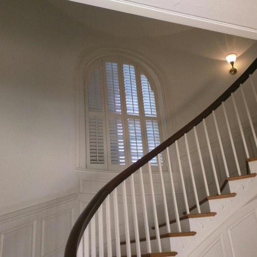 White plantation shutters covering arched window located in round stairwell.